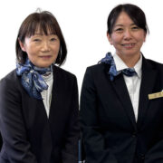 Two women in black suits and blue scarves posing against a white background, one with a name badge on her lapel (professional staff photo).